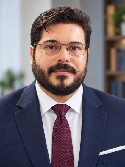 Retrato de Valber Gregory, de terno azul-marinho e gravata vinho, em um escritório com estantes ao fundo.