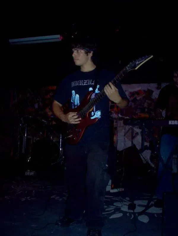 Valber, jovem, de camiseta preta, tocando uma guitarra vermelha em um palco escuro, com bateria e teclado ao fundo.