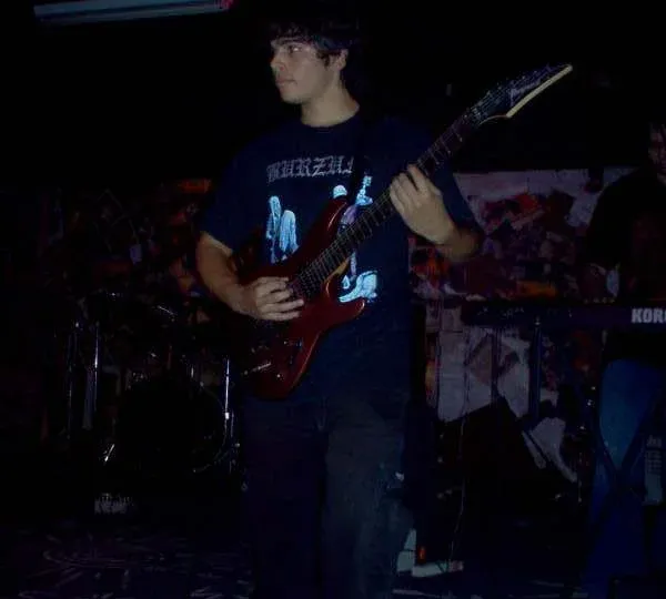 Valber, jovem, de camiseta preta, tocando uma guitarra vermelha em um palco escuro, com bateria e teclado ao fundo.