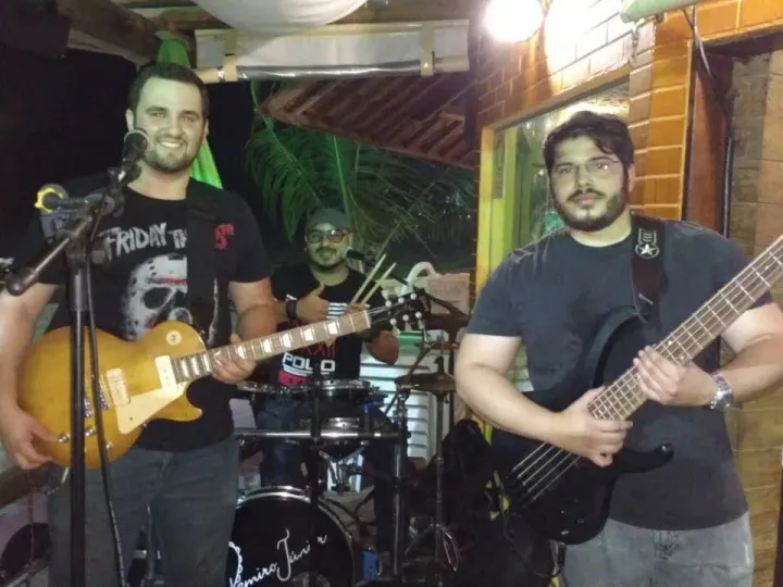 Três músicos em um bar ao ar livre à noite: vocalista com guitarra Les Paul ao centro, baterista atrás e Valber com baixo de cinco cordas à direita.