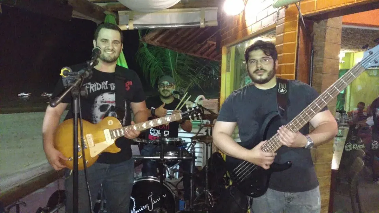 Três músicos em um bar ao ar livre à noite: vocalista com guitarra Les Paul ao centro, baterista atrás e Valber com baixo de cinco cordas à direita.