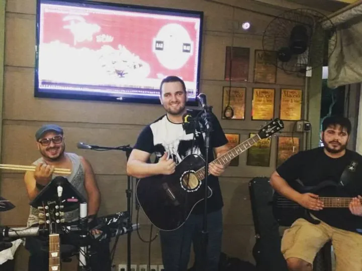 Trio tocando em um bar: baterista com baquetas erguidas, vocalista com violão ao centro e Valber sentado com o baixo, sob uma TV e quadros na parede.