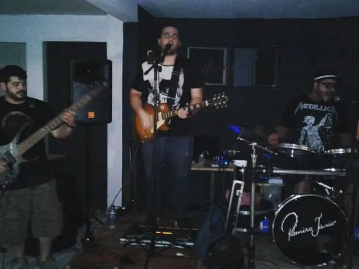 Trio em apresentação em um salão: Valber ao baixo à esquerda, vocalista com guitarra ao centro e baterista à direita, com o nome Ramiro Júnior no bumbo.