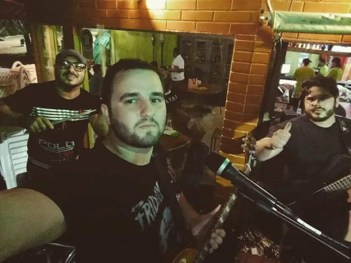 Selfie do vocalista com o baterista e Valber, que faz sinal de positivo com o baixo, em um bar de tijolos aparentes.