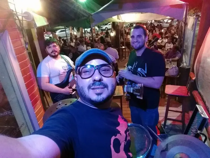 Selfie do baterista, de boina e óculos, com o vocalista e Valber ao fundo, em um bar cheio à noite.