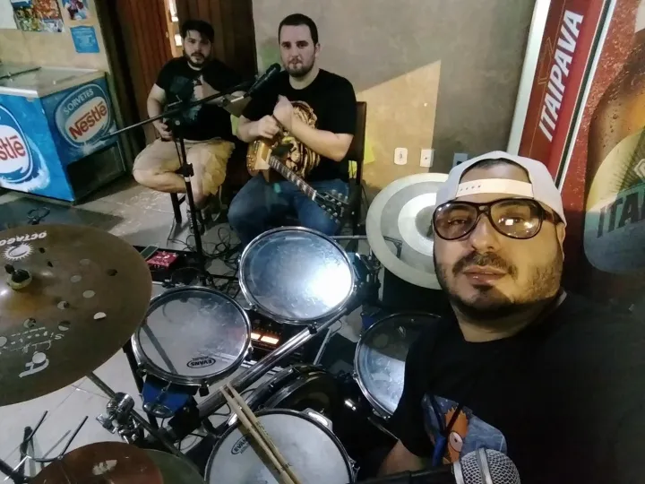 Selfie do baterista atrás da bateria; ao fundo, Valber e o vocalista sentados com os instrumentos.
