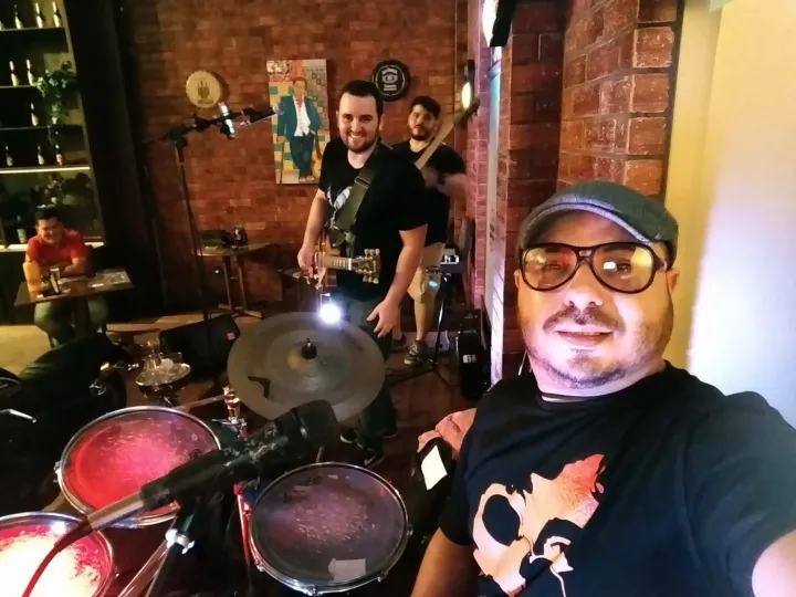 Selfie do baterista em um bar de tijolos; ao fundo, o vocalista com a guitarra e Valber com o baixo.