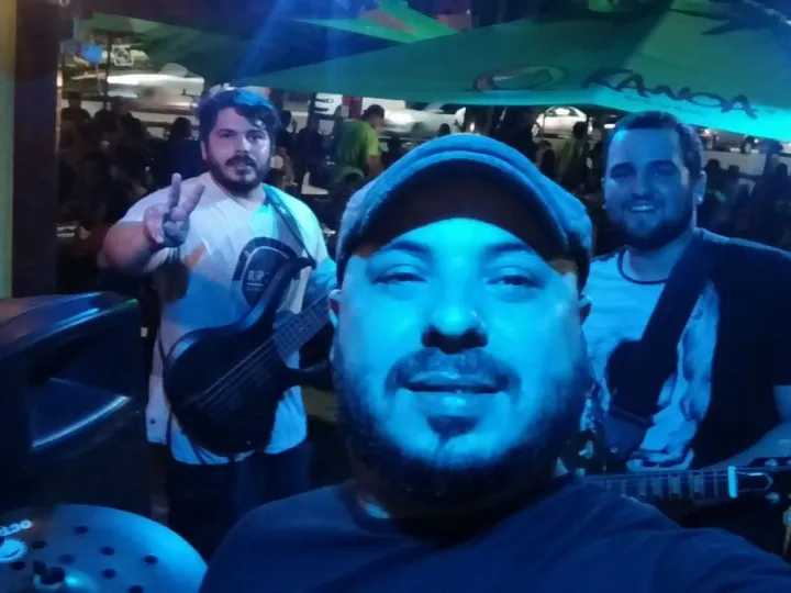 Selfie do baterista, de boné, com Valber ao baixo e o vocalista ao fundo, sob guarda-sóis de um bar à noite, com luz azul.