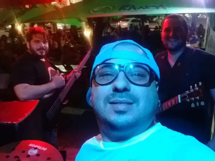 Selfie do baterista com luz azul; ao fundo, Valber faz sinal de paz com o baixo e o vocalista sorri com a guitarra.