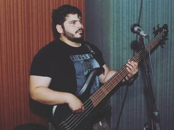 Valber tocando um baixo elétrico de cinco cordas, de pé, diante de um microfone, com parede vermelha ao fundo.