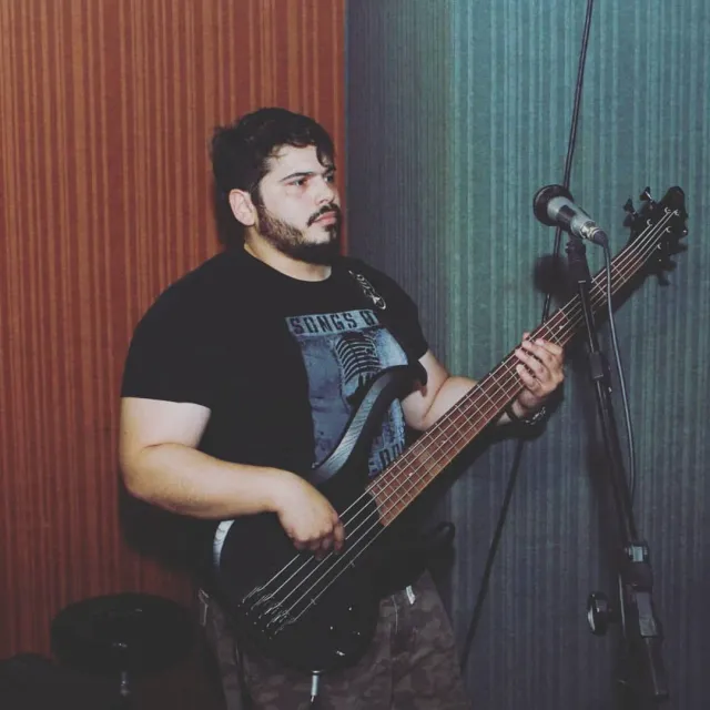 Valber tocando um baixo elétrico de cinco cordas, de pé, diante de um microfone.